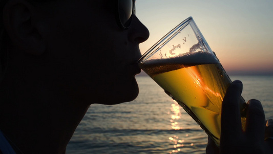 日落时一位戴太阳镜的年轻女子在海滩上喝啤酒视频