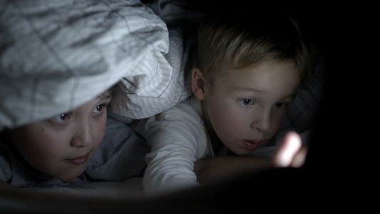 两个男孩晚上躺在床上的毯子下在触摸板上看卡通或电影视频