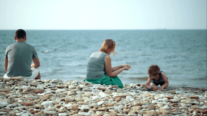三口之家在卵石滩上母子玩得很开心父亲静静地坐着看着大海55秒视频