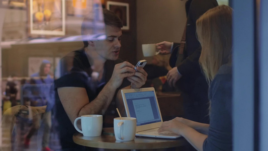 通过玻璃拍摄咖啡馆里的年轻夫妇丈夫在看着智能手机妻子在用笔记本电脑工作视频