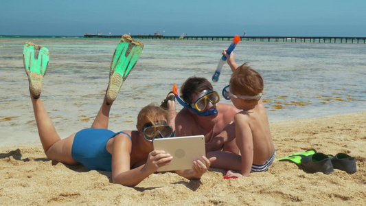 潜水后父母和儿子躺在海滩上他们在平板电脑上看东西说话视频