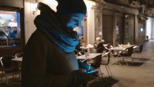 在夜间小街的户外咖啡馆附近一个戴着帽子的女人在用智能手机视频