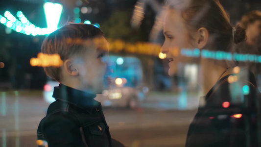 透过玻璃窗拍摄夜晚年轻母子在繁忙的街道上小男孩兴奋地和妈妈说话视频