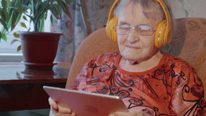 老年妇女在家里坐在扶手椅上用平板电脑和无线耳机听音乐19秒视频