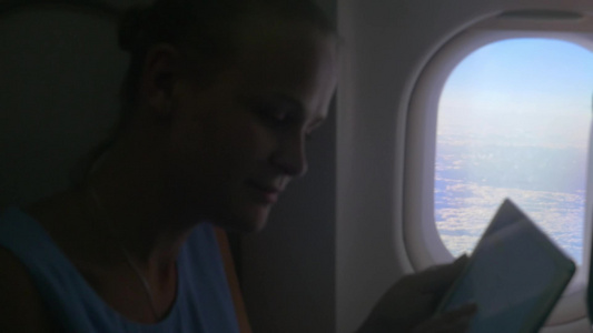 一个年轻的女人坐在飞机窗口在用平板电脑打字视频