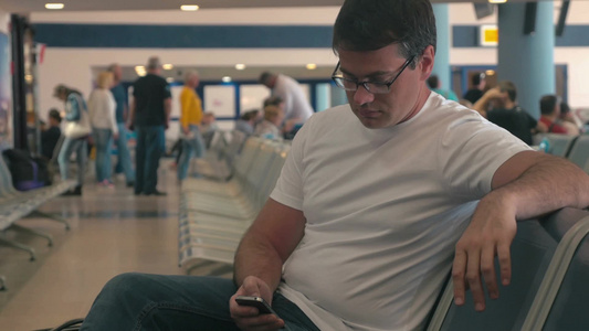 年轻人戴着眼镜在机场或车站的候车室里拿着手机上网浏览信息视频