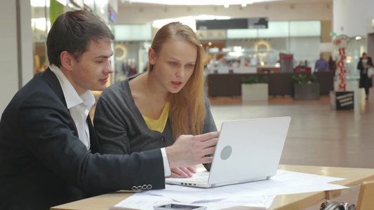 两个商人坐在商场休息区用笔记本电脑讨论他们的新项目视频