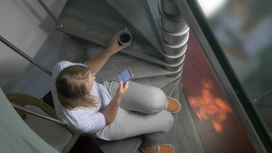 一位年轻女子坐在螺旋楼梯上喝着咖啡玩智能手机视频