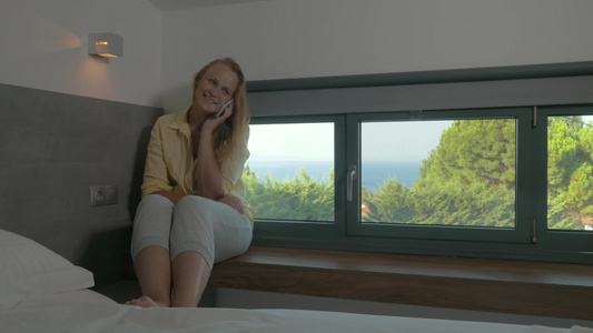 坐在酒店房间的窗台上年轻漂亮的女人在电话交谈视频