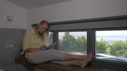 年轻的微笑的女人坐在窗台上玩平板电脑视频