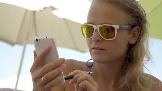 年轻微笑的金发女人在炎热的海滩玩手机视频