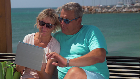 年长的夫妇戴着太阳镜坐在海滨的海滩上使用平板电脑上浏览网页视频