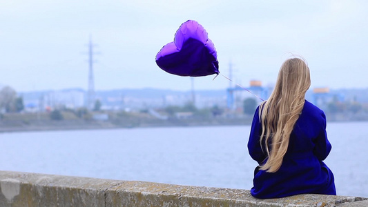 年轻女子穿钴蓝色大衣手里拿着蓝色爱心气球独自坐在河堤的栏杆上视频
