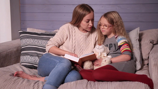 年轻迷人的妈妈和可爱的小女孩坐在家里的沙发上一起读故事视频