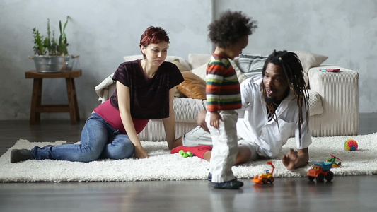 美丽的白种人怀孕母亲和黑种人丈夫一起陪可爱的蹒跚学步的儿子坐在客厅的地板上玩玩具视频