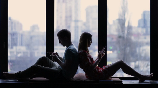 年轻夫妇背靠背坐在家里飘窗上玩手机视频