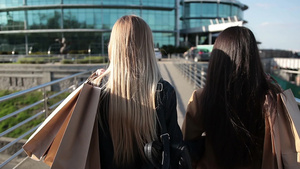 后景留着长发的两个美丽的女人肩上拿着购物袋在日落时漫步在人行桥上。 14秒视频
