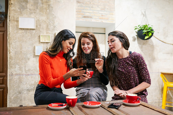 由三个快乐的女性朋友成的多民族群体咖啡馆酒吧里看着智能手机三个快乐的朋友咖啡馆里喝咖啡图片