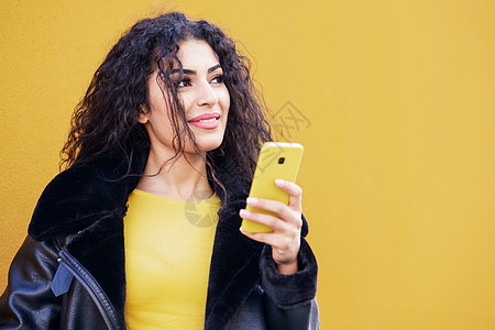 年轻的阿拉伯女人穿着休闲服,用她的智能手机黄色的城市墙上录制语音音符阿拉伯女人用她的智能手机走街上图片