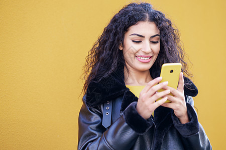 年轻的阿拉伯女人用智能手机黄色的城市墙上发短信年轻的阿拉伯女人用她的智能手机走街上图片
