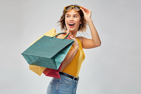 销售人们的快乐的微笑年轻妇女芥末黄色上衣牛仔裤与购物袋灰色背景带着购物袋的快乐微笑的年轻女人图片