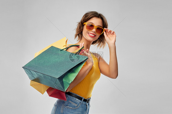 销售人们的快乐的微笑年轻妇女芥末黄色上衣牛仔裤与购物袋灰色背景带着购物袋的快乐微笑的年轻女人图片