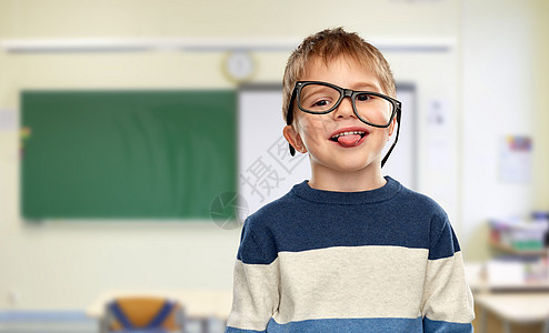 学校教育视觉微笑的小男孩的肖像,戴着歪歪扭扭的眼镜,教室背景上露出舌头戴眼镜的小男孩学校表现出舌头图片
