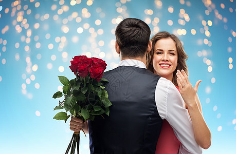爱情,夫妻,求婚人的快乐的女人与订婚戒指束玫瑰拥抱男人节日灯上的蓝色背景女人带着订婚戒指玫瑰拥抱男人图片