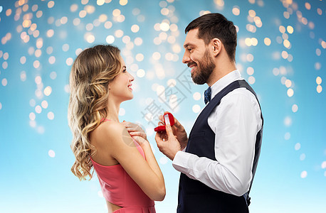 爱情,夫妻,求婚人的快乐的男人小红盒子里给女人钻石订婚戒指,蓝色背景的节日灯上男人情人节给女人订婚戒指图片