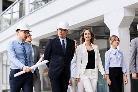 建筑建筑商业快乐的建筑师小戴着头盔,蓝图沿着办公楼走戴头盔的商业队沿着办公室走图片