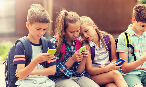 小学教育,友谊,童年,技术人的群快乐的小学生,智能手机背包坐户外长凳上有智能手机的小学生图片