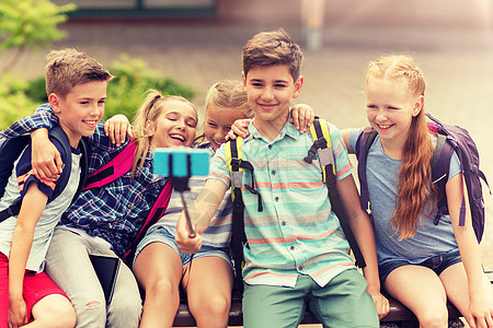 小学教育,技术,友谊,童年人的群小学生着背包坐长凳上,用智能手机户外自拍杆上拍照快乐的小学生自拍图片