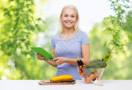 健康的饮食,烹饪,食物,技术人的快乐的微笑年轻妇女与平板电脑碗蔬菜绿色的自然背景微笑的年轻女人用平板电脑做饭图片