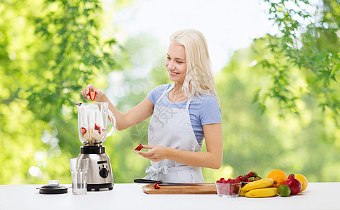 健康的饮食,烹饪,食物人的快乐的微笑年轻妇女,把水果浆果水果冰沙搅拌机绿色的自然背景微笑的女人用搅拌机做水果奶昔图片