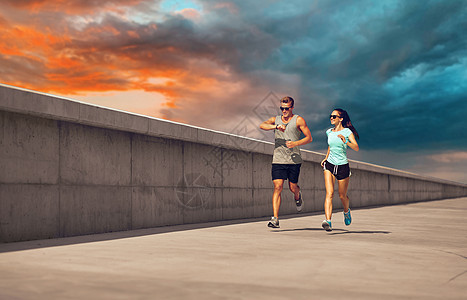 健身,运动生活方式的幸福的夫妇穿着运动服装太阳镜沿着混凝土码头运行日落天空的背景穿着运动服的夫妇沿着码头跑步图片