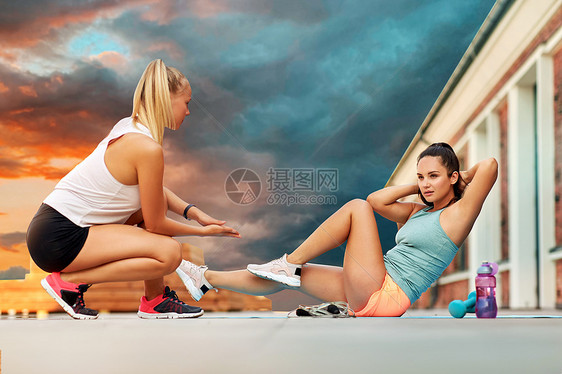 健身,运动健康的生活方式年轻的女人帮助她的朋友屋顶上的垫子上做自行车皱褶,日落的天空背景上妇女户外训练做自行图片
