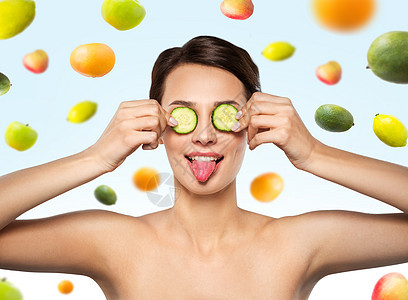 美丽,护肤排美丽的女人制作黄瓜眼罩,并蓝色背景的水果上展示她的舌头美丽的女人用黄瓜做眼罩图片