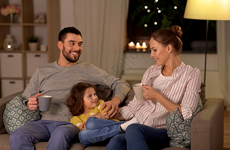 家庭人的幸福微笑的父亲,母亲喝茶,小女儿晚上坐家里的沙发上幸福家庭坐家里沙发上的肖像图片