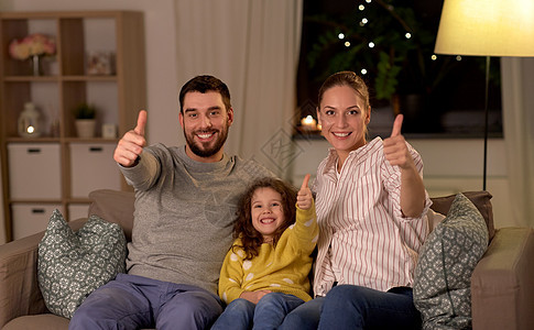 家庭人的幸福微笑的父亲,母亲小女儿坐沙发上,晚上家里竖起大拇指幸福家庭坐家里沙发上的肖像图片