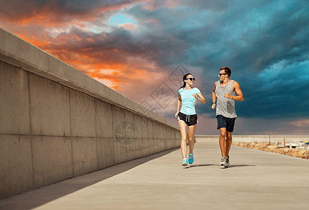 健身,运动人的幸福的夫妇穿着运动服装太阳镜沿着码头日落的天空背景上运行穿着运动服的夫妇沿着码头跑步图片