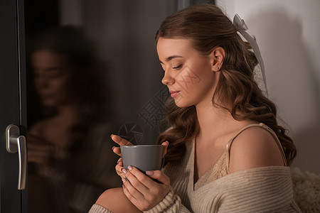 冬天,舒适人的年轻的女人穿着套头衫,坐窗户上,家喝咖啡或茶杯家里窗户上有咖啡或茶杯的女人图片