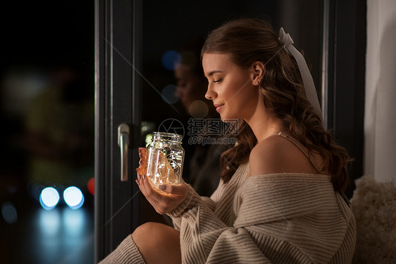 冬天,舒适人的年轻的女人穿着套衫,坐窗户上,玻璃石匠罐子杯里装着花环家里的玻璃杯里有花环灯的女人图片
