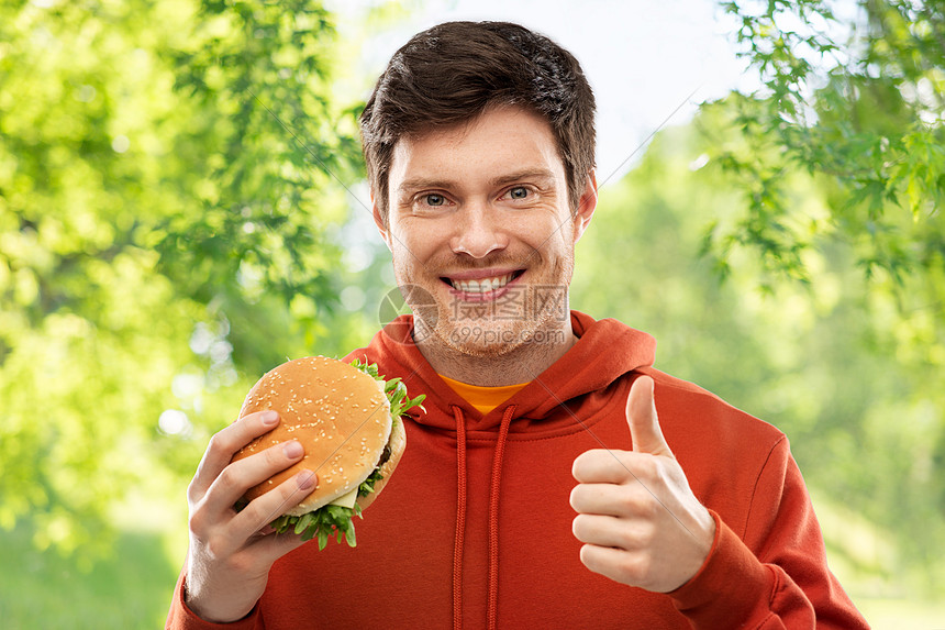 快餐,健康的饮食人们的快乐的微笑年轻人穿着红色连帽衫,汉堡包大拇指绿色的自然背景上快乐的年轻人用汉堡包竖起大图片