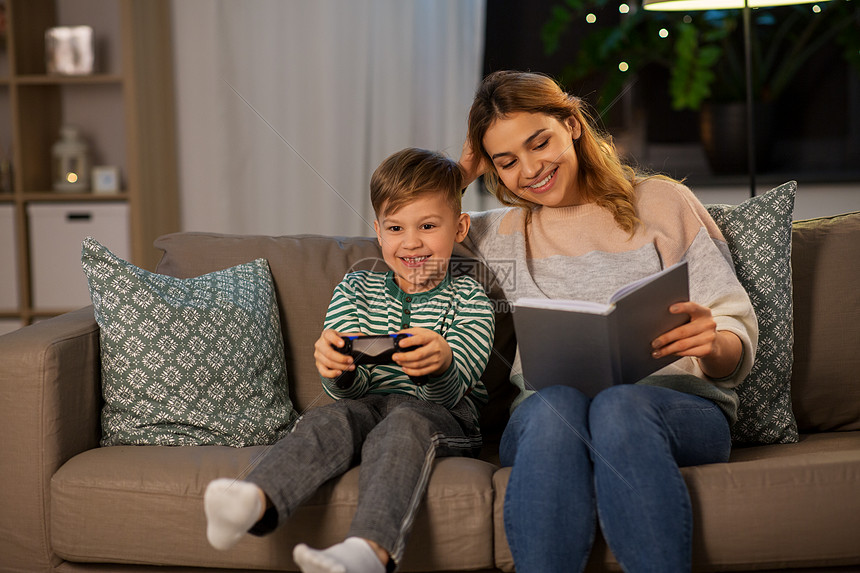 家庭,休闲人的快乐微笑的母亲阅读书她的小儿子与游戏垫晚上玩电子游戏快乐的母亲小儿子家里度过时光图片