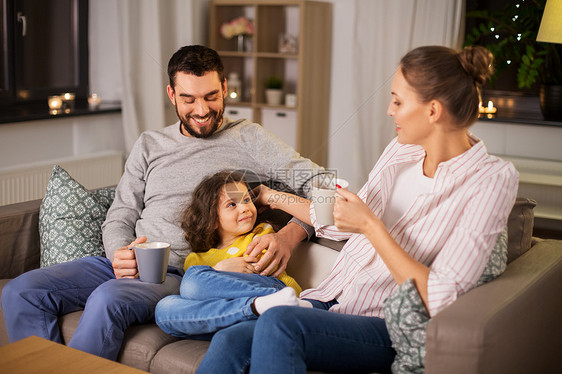 家庭人的幸福微笑的父亲,母亲喝茶,小女儿晚上坐家里的沙发上幸福家庭坐家里沙发上的肖像图片