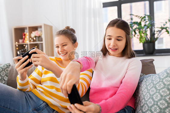 人,技术友谊的快乐的十几岁的女孩坐沙发上玩游戏家女孩们家里用智能手机玩游戏图片