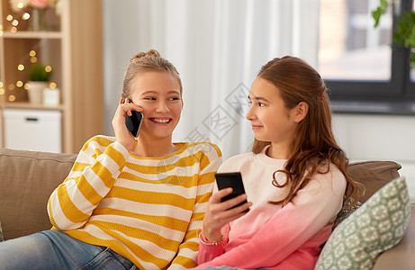 彩沙人,技术友谊的快乐的十几岁的女孩坐沙发上的智能手机家快乐的十几岁的女孩家里有智能手机背景