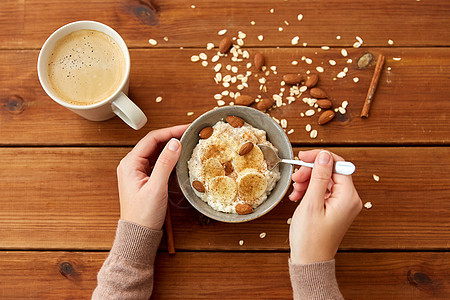 食物早餐妇女的手吃燕麦粥碗与切片香蕉,杏仁坚果肉桂杯咖啡木桌上手燕麦片早餐杯咖啡图片