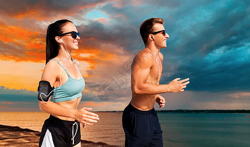 健身,运动健康的生活方式快乐的夫妇与耳机手臂乐队运行海上日落天空背景夫妇带着电话手臂乐队海滩上跑步图片