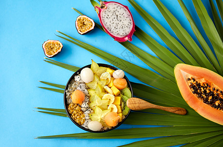 芒果火龙果食物,夏天健康的饮食混合同的异国水果碗与木勺绿色风扇棕榈叶蓝色背景用木勺把异国水果混合碗里背景